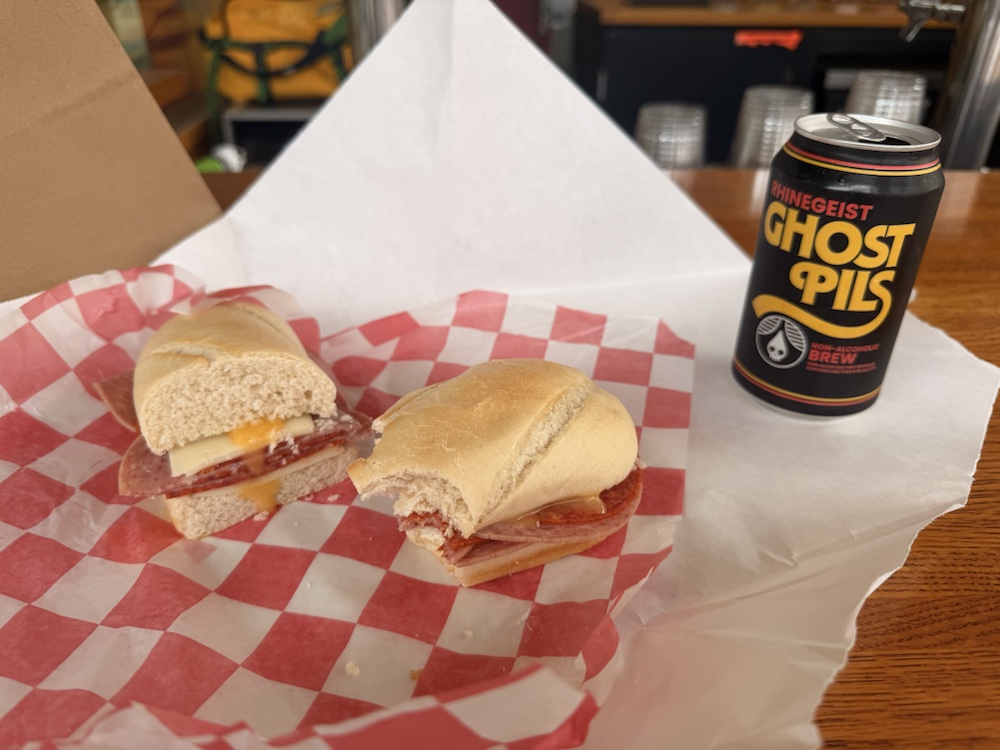 A sandwich on deli paper beside a can of Rhinegeist Ghost Pils at a wooden table.